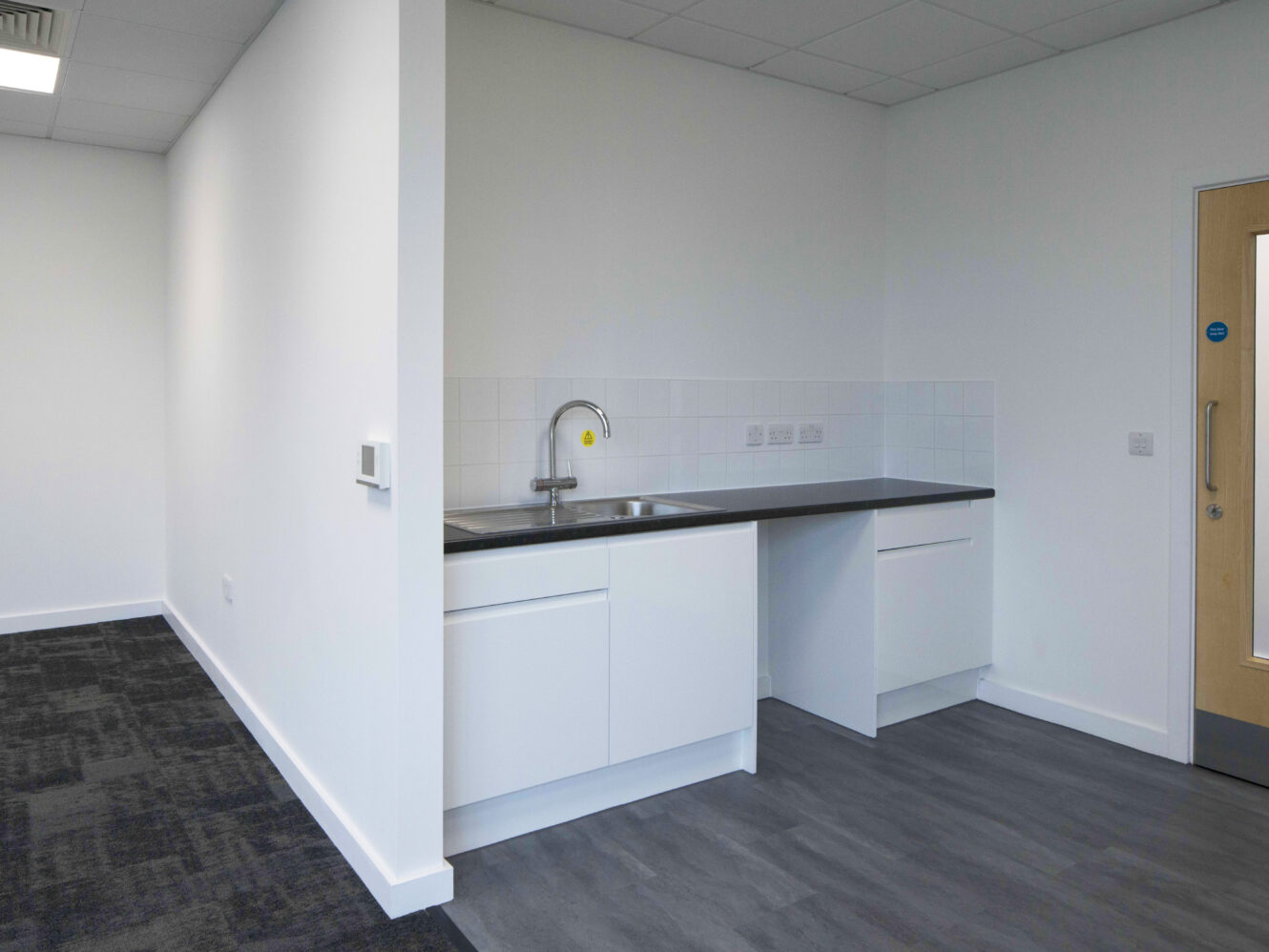 A small, modern kitchenette with white cabinets, a stainless steel sink, and dark countertops, adjacent to a door and an office area with dark carpet tiles.