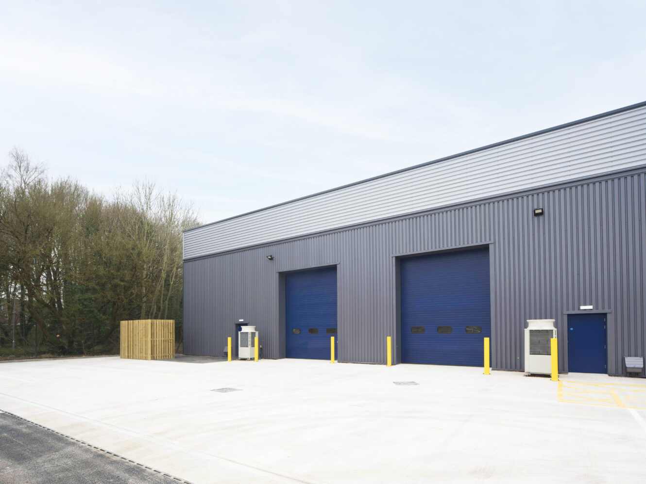 Gray industrial warehouse with two large blue roller doors, a small blue entrance door, yellow safety bollards, and trees in the background.