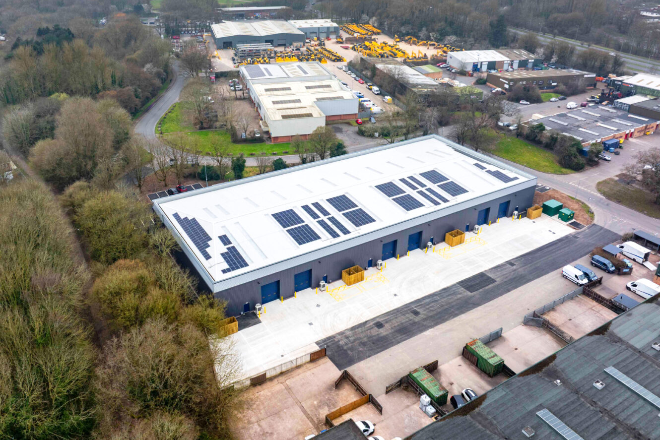 Aerial view of an industrial warehouse with solar panels on the roof, surrounded by trees, roads, and other industrial buildings.
