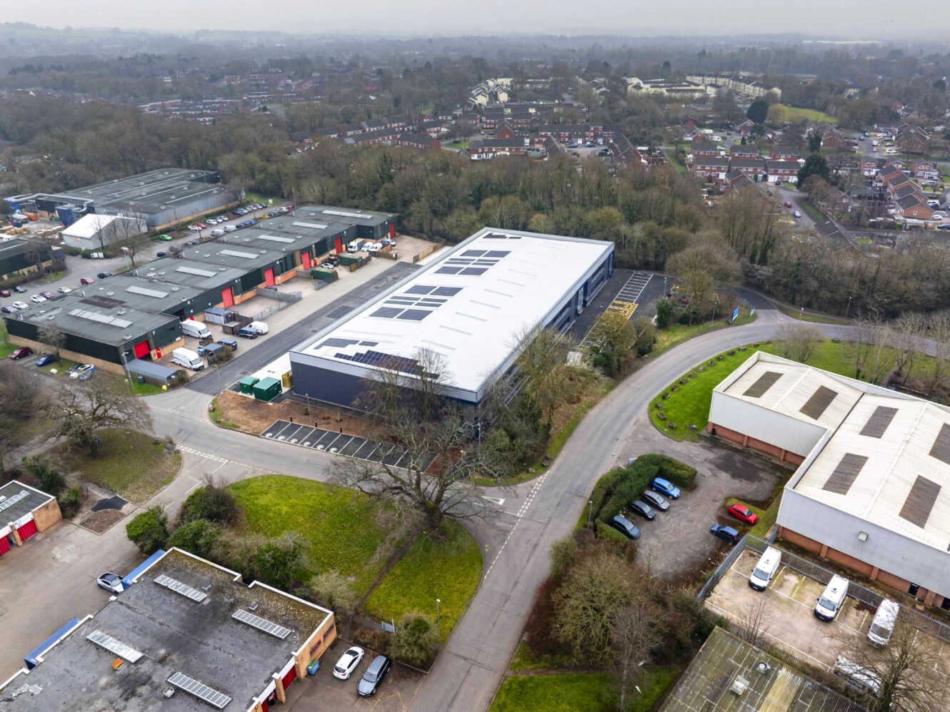 Aerial view of an industrial estate with warehouses, parked vehicles, surrounding roads, and nearby residential houses bordered by trees.