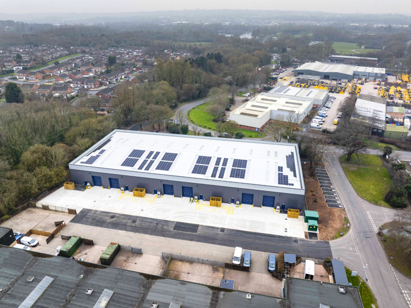 Aerial view of a large industrial warehouse with solar panels on its roof, surrounded by trees, other buildings, and a residential area.