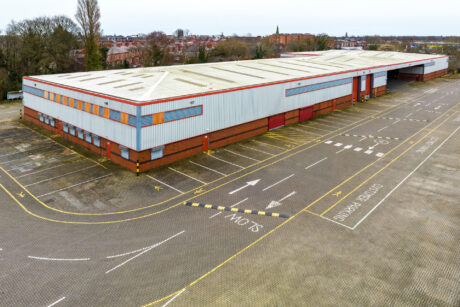 A large, empty warehouse with a brick and metal exterior is surrounded by an expansive, vacant parking lot marked with lanes and traffic instructions.