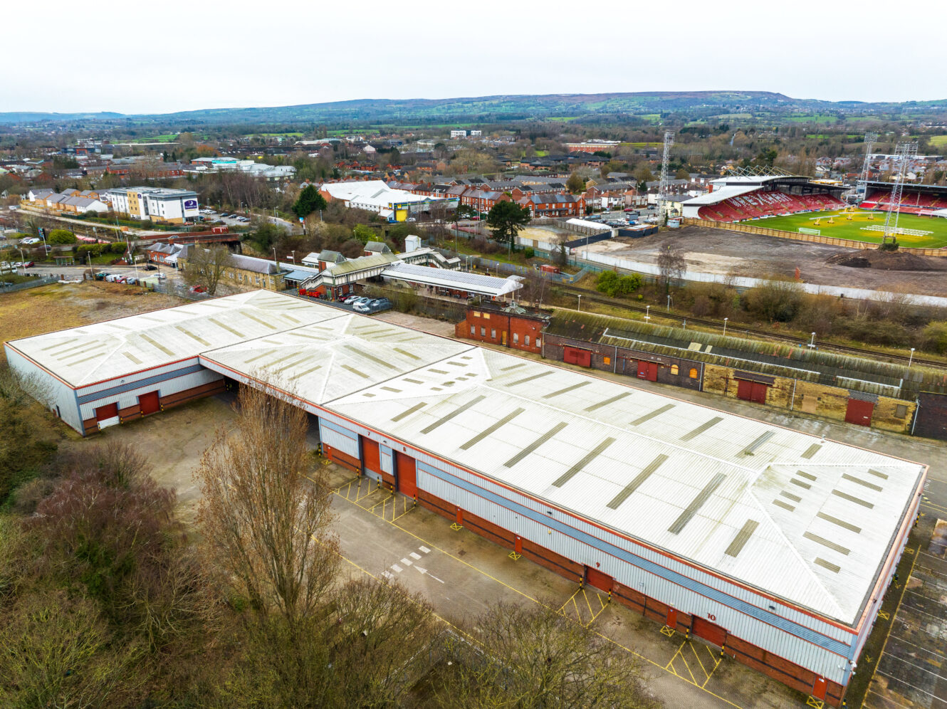 A large industrial warehouse with a white roof is surrounded by trees, parking lots, and nearby sports fields in a suburban area.
