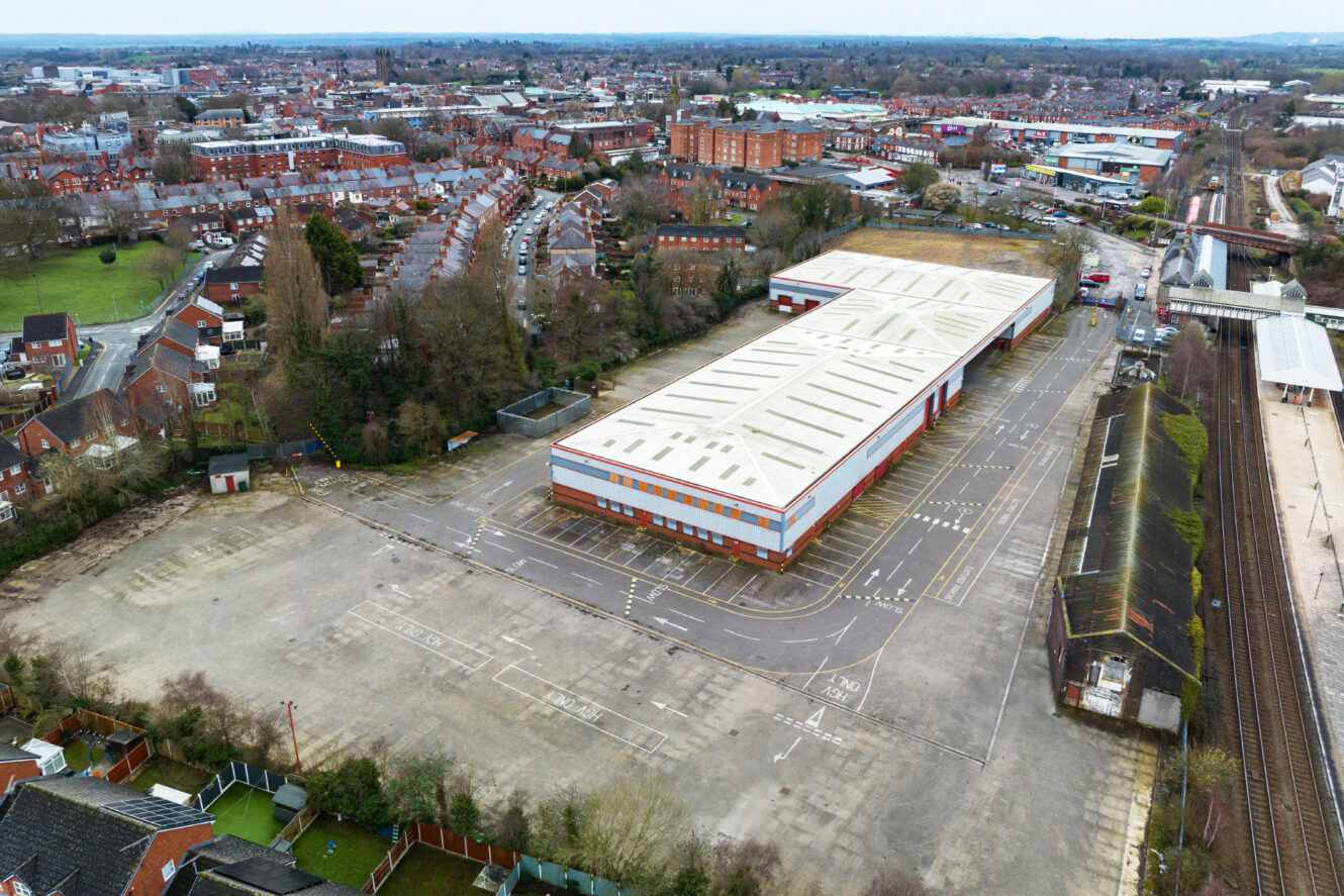 Aerial view of a large, mostly empty parking lot surrounding a warehouse-style building, with residential and commercial areas in the background.