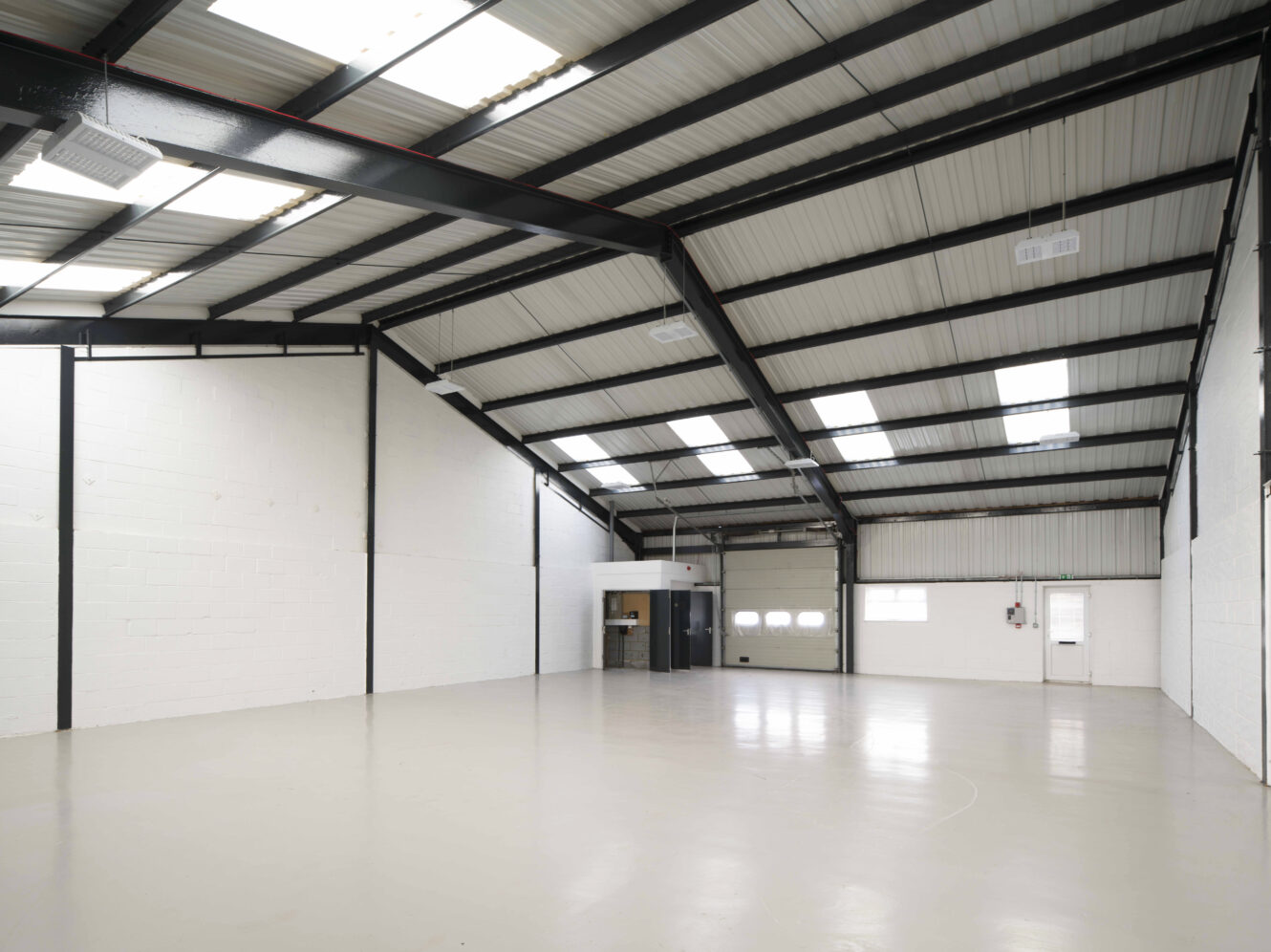 Interior of an empty industrial warehouse with white walls, high metal ceiling, polished concrete floor, and a large garage door at the far end.