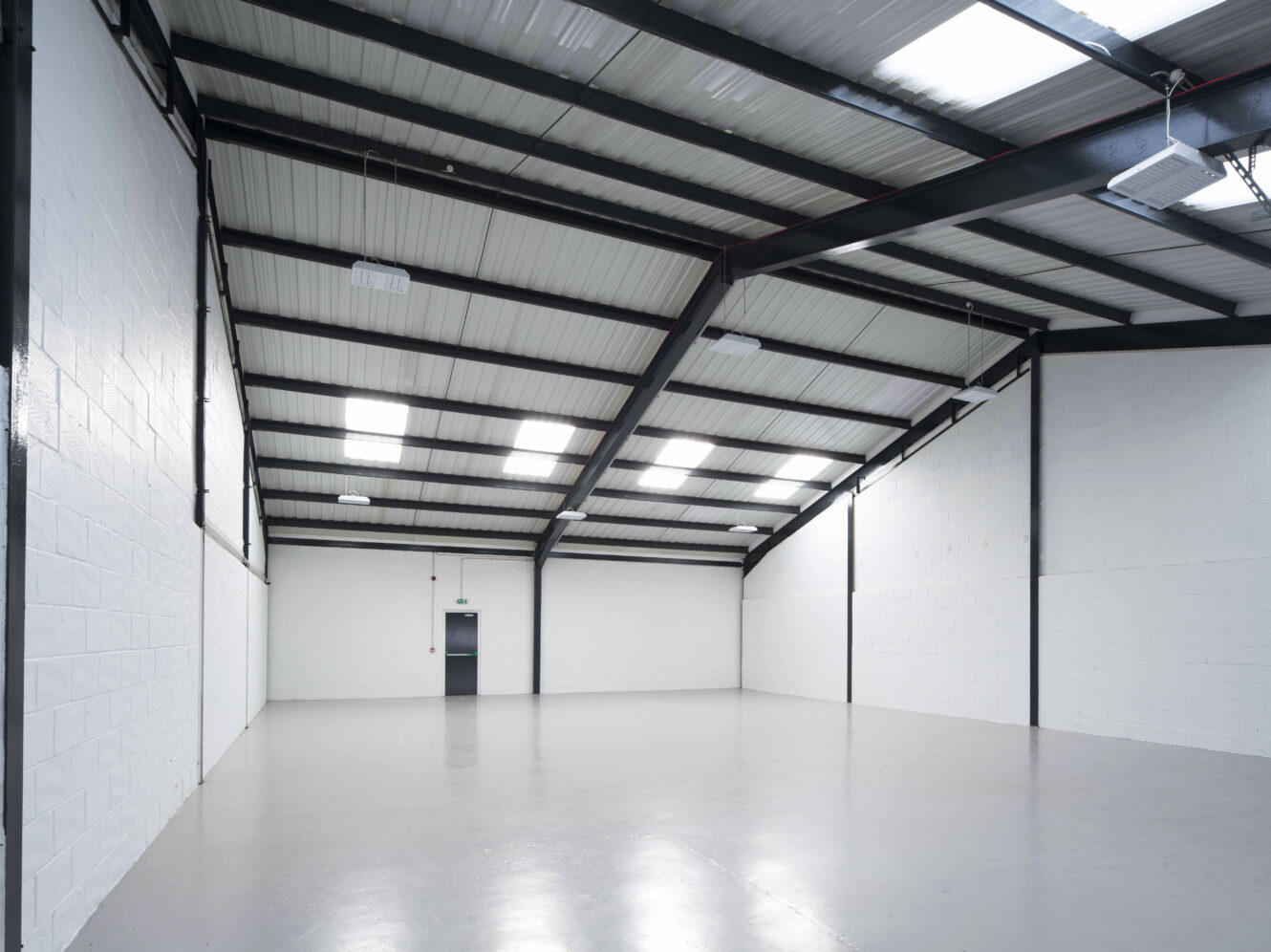 Empty industrial warehouse with white walls, a gray polished floor, black steel beams, and skylights in the ceiling. A single door is visible at the far end.
