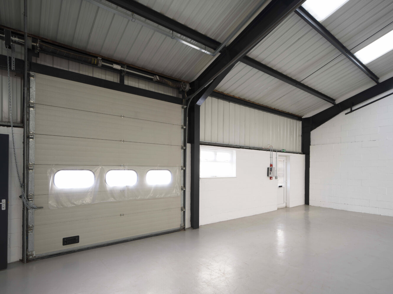 Empty industrial warehouse interior with a large roller door, white painted brick walls, a small window, and a white exit door with emergency equipment nearby.