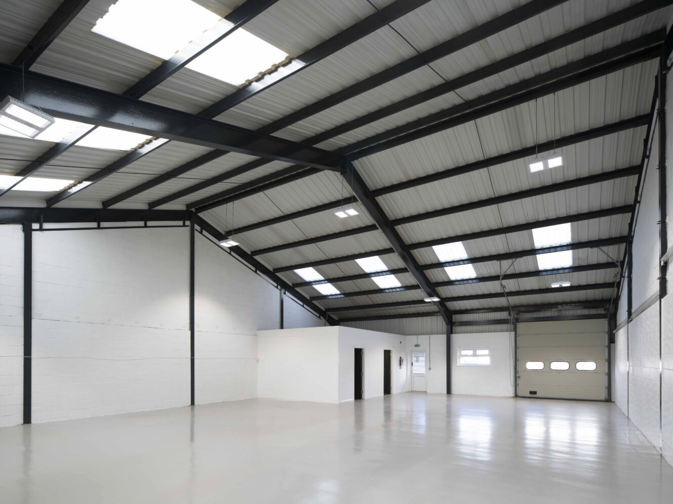 Spacious, empty warehouse interior with white walls, high ceiling, skylights, and a large roller door. The floor is clean and polished.
