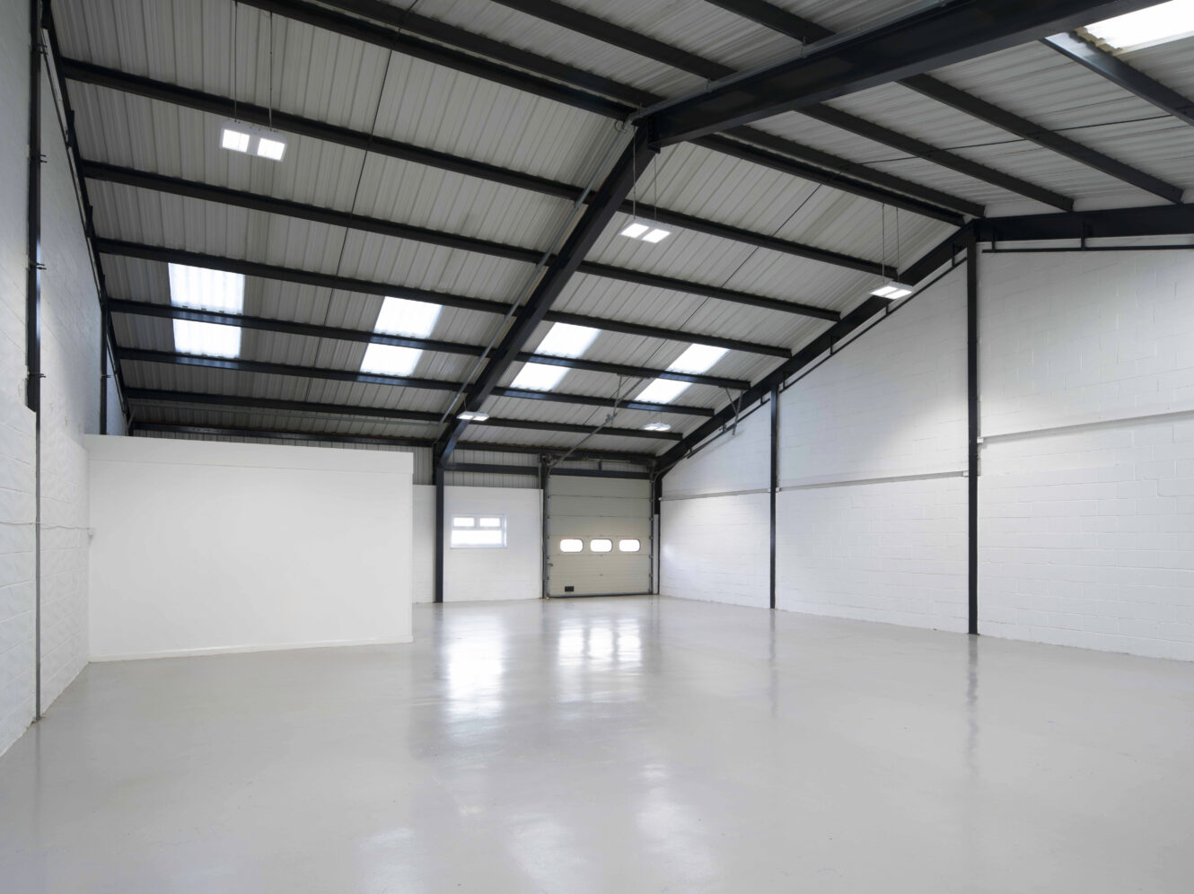 Interior of an empty industrial warehouse with white walls, high ceiling, skylights, and a large closed garage door at the far end.