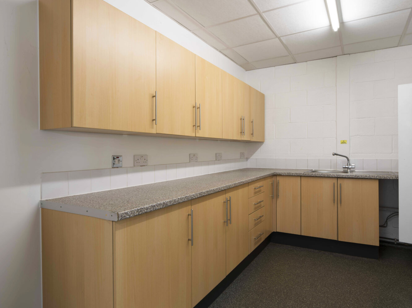 A simple kitchen with wooden cabinets, granite countertops, a sink, and a fluorescent ceiling light. The walls are white with visible power outlets.