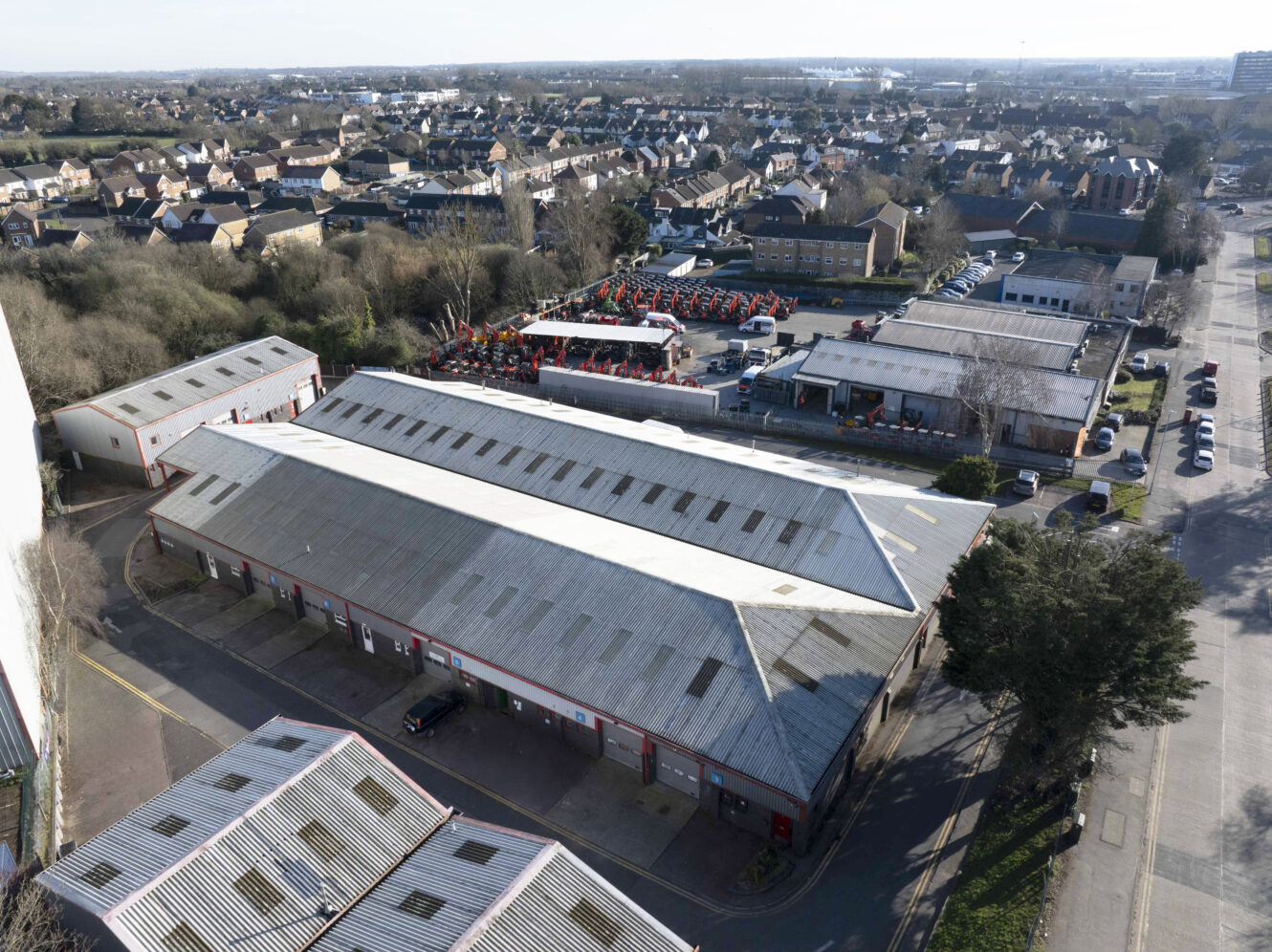 Aerial view of an industrial warehouse complex with parking area, surrounded by residential houses and trees in a suburban setting.