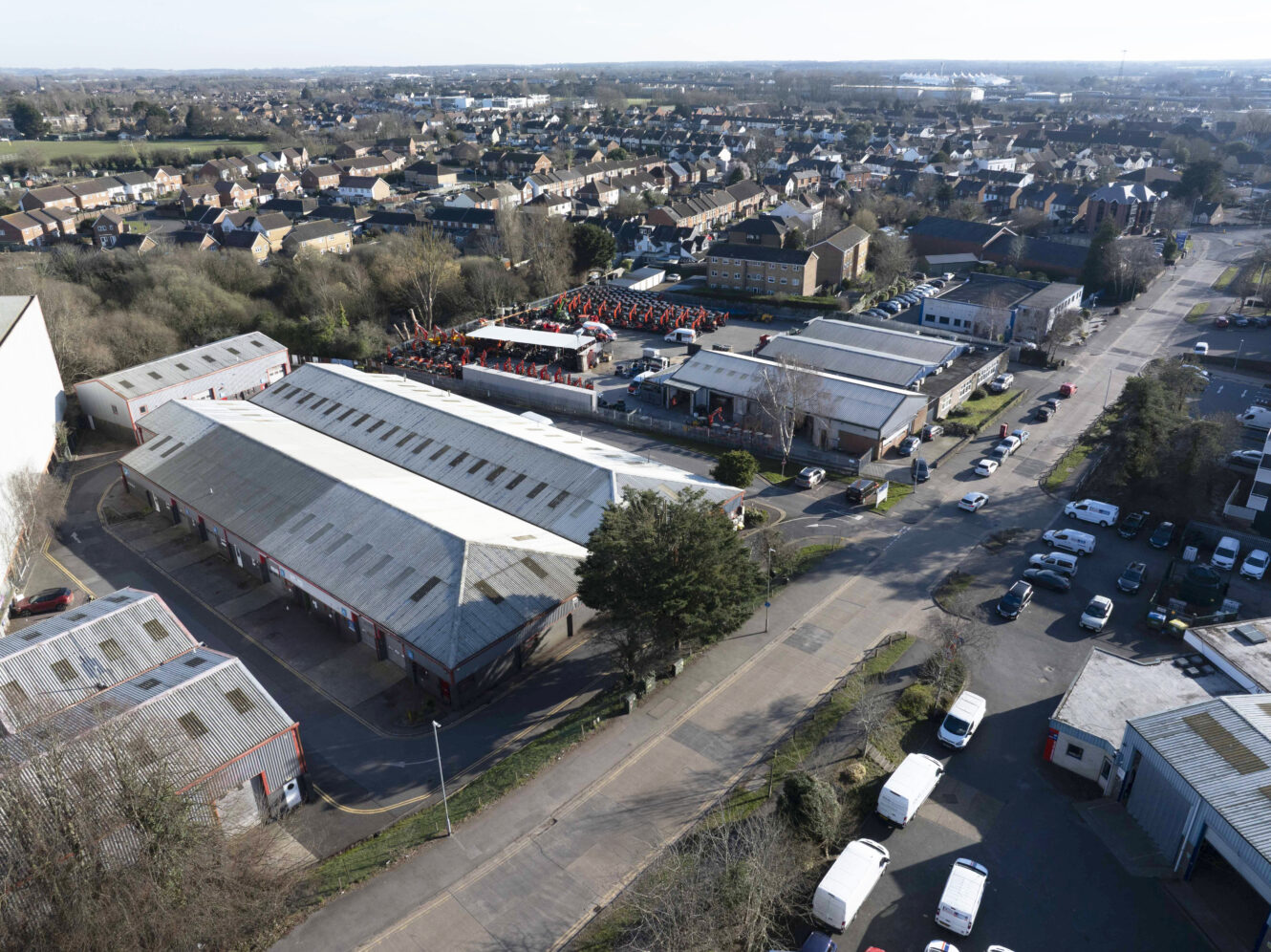 Aerial view of an industrial area with large warehouses, parked vehicles, and adjacent residential neighborhood in the background.