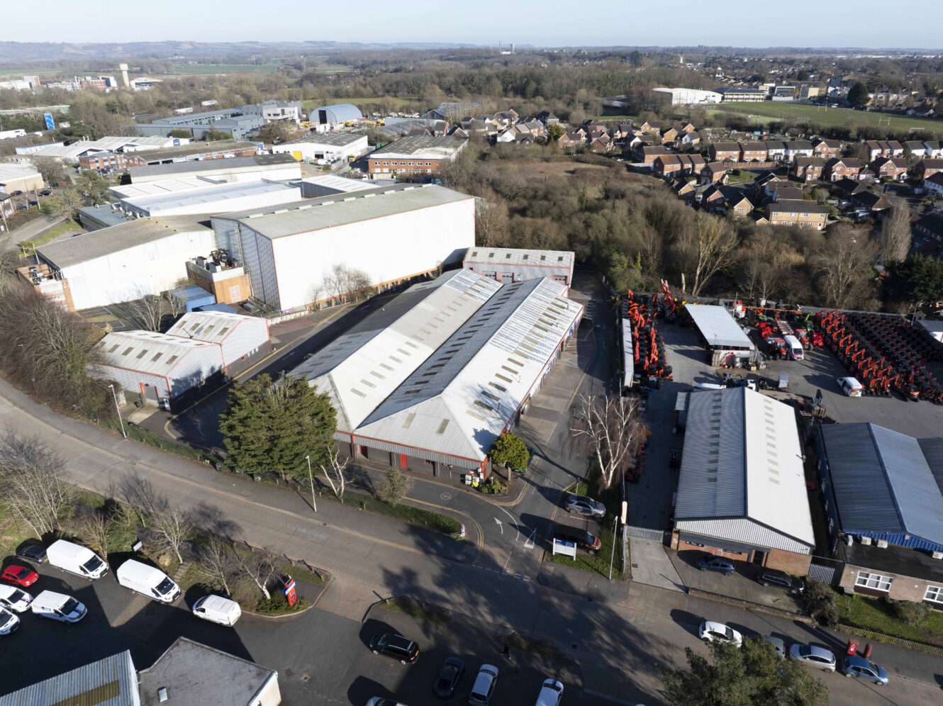 Aerial view of an industrial area with warehouses, parked vehicles, and nearby residential houses, surrounded by trees and roads.