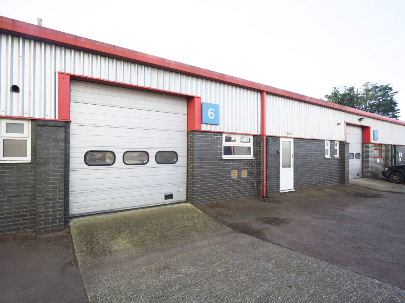 A row of industrial units with brick and metal exteriors, showing unit 6 with a closed roller shutter door, a white door, and numbered blue signs above each unit.