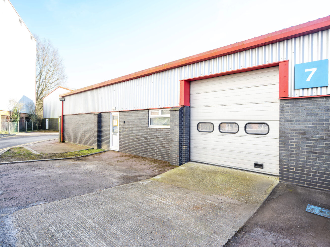Single-story industrial unit with a gray brick facade, metal siding, a white overhead garage door, and a blue sign displaying the number 7.