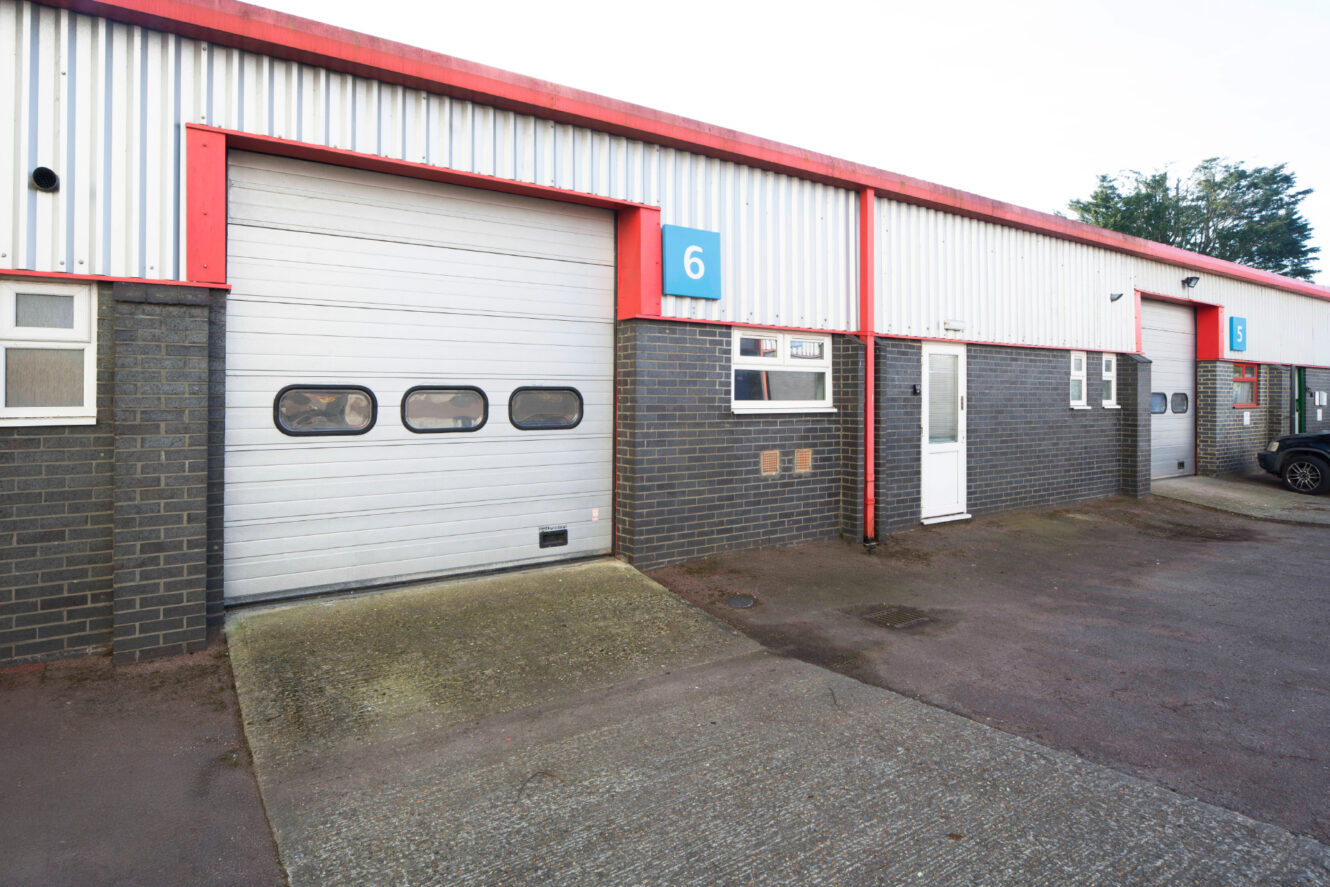 Exterior of an industrial unit with a large roller shutter door, a white entry door, and the number 6 sign above the entrance.