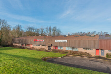 A large, single-story industrial building with Honda and Thunder Road signs on the facade, surrounded by grass and trees under a clear blue sky.