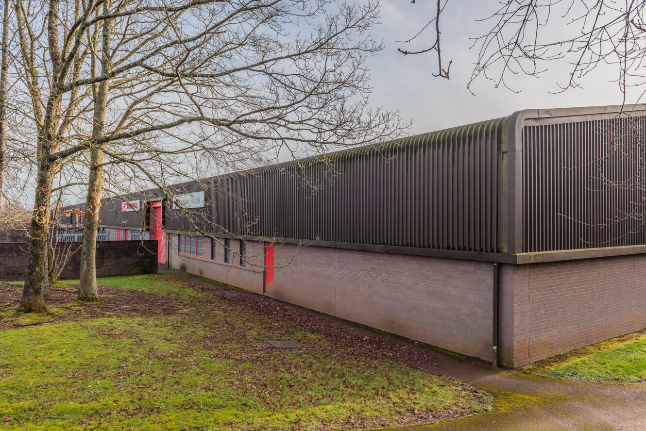 A low, modern building with dark metal siding and red accents, situated next to leafless trees and a grassy area under a cloudy sky.