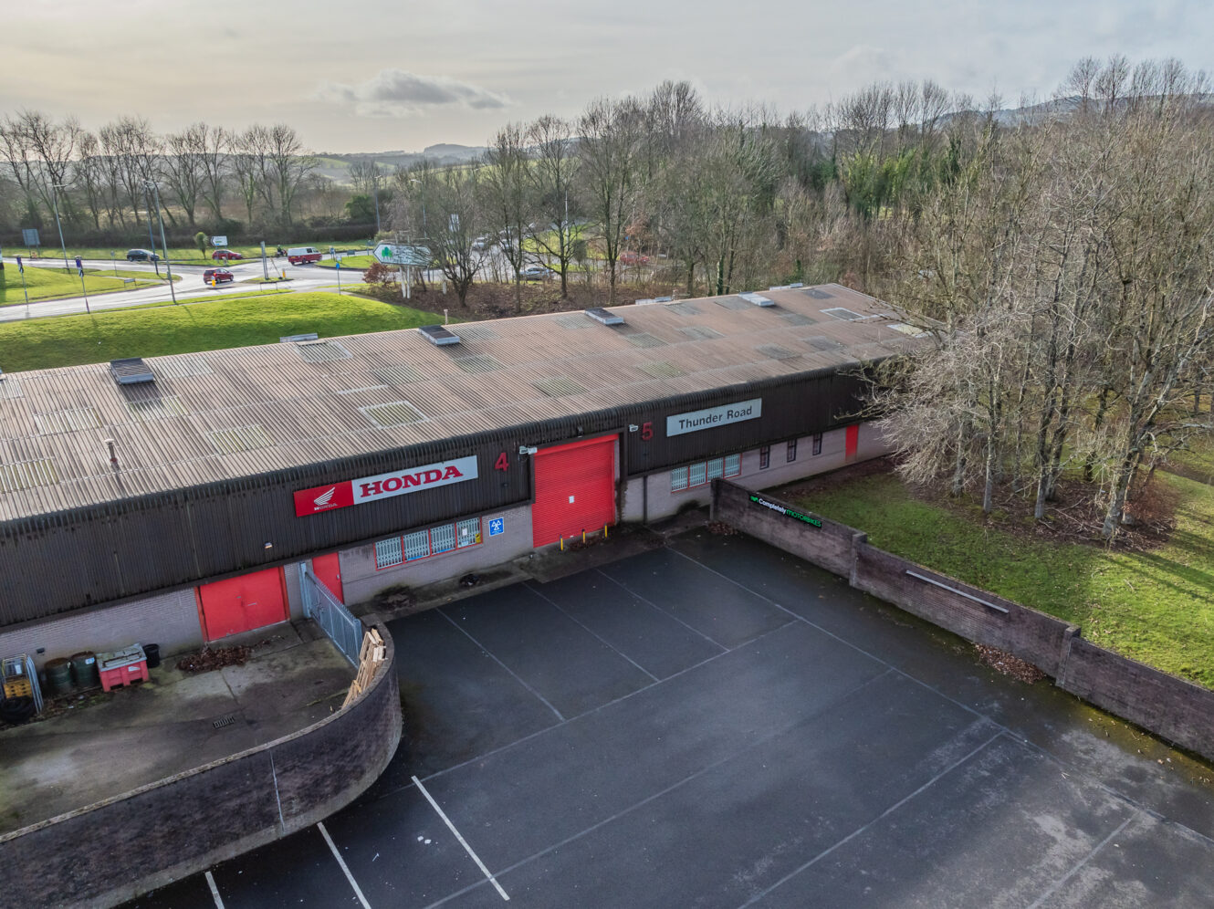A large industrial building with Honda signage, red doors, and an empty parking lot, surrounded by trees and a road in the background.