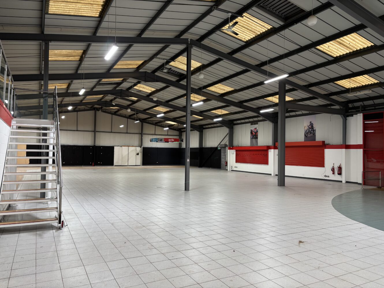 Large empty industrial warehouse with tiled floor, metal ceiling, fluorescent lighting, a staircase on the left, and red accents on some walls.