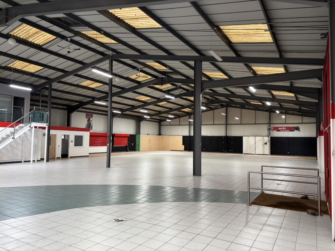 Spacious industrial interior with tiled flooring, metal roof, exposed beams, and a small mezzanine area; walls are red, white, and black.