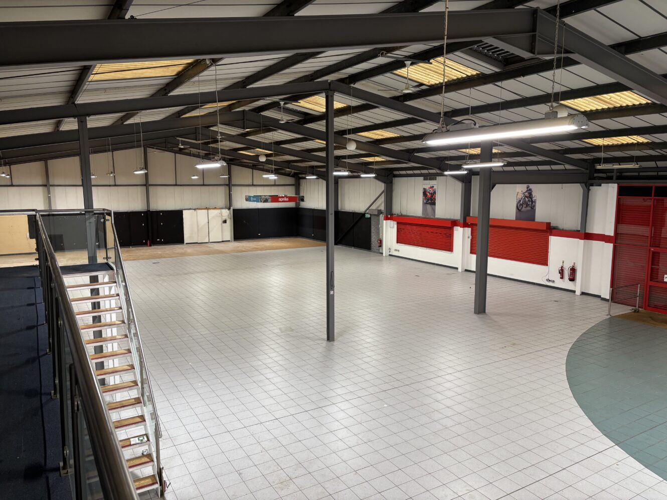 Spacious, empty industrial room with tiled floor, exposed ceiling beams, staircase on the left, and red and white walls with a few posters and fire extinguishers.