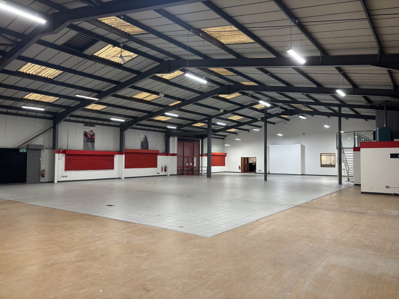 Spacious, well-lit industrial warehouse with tiled and wooden flooring, red and white walls, exposed steel beams, and high ceilings.