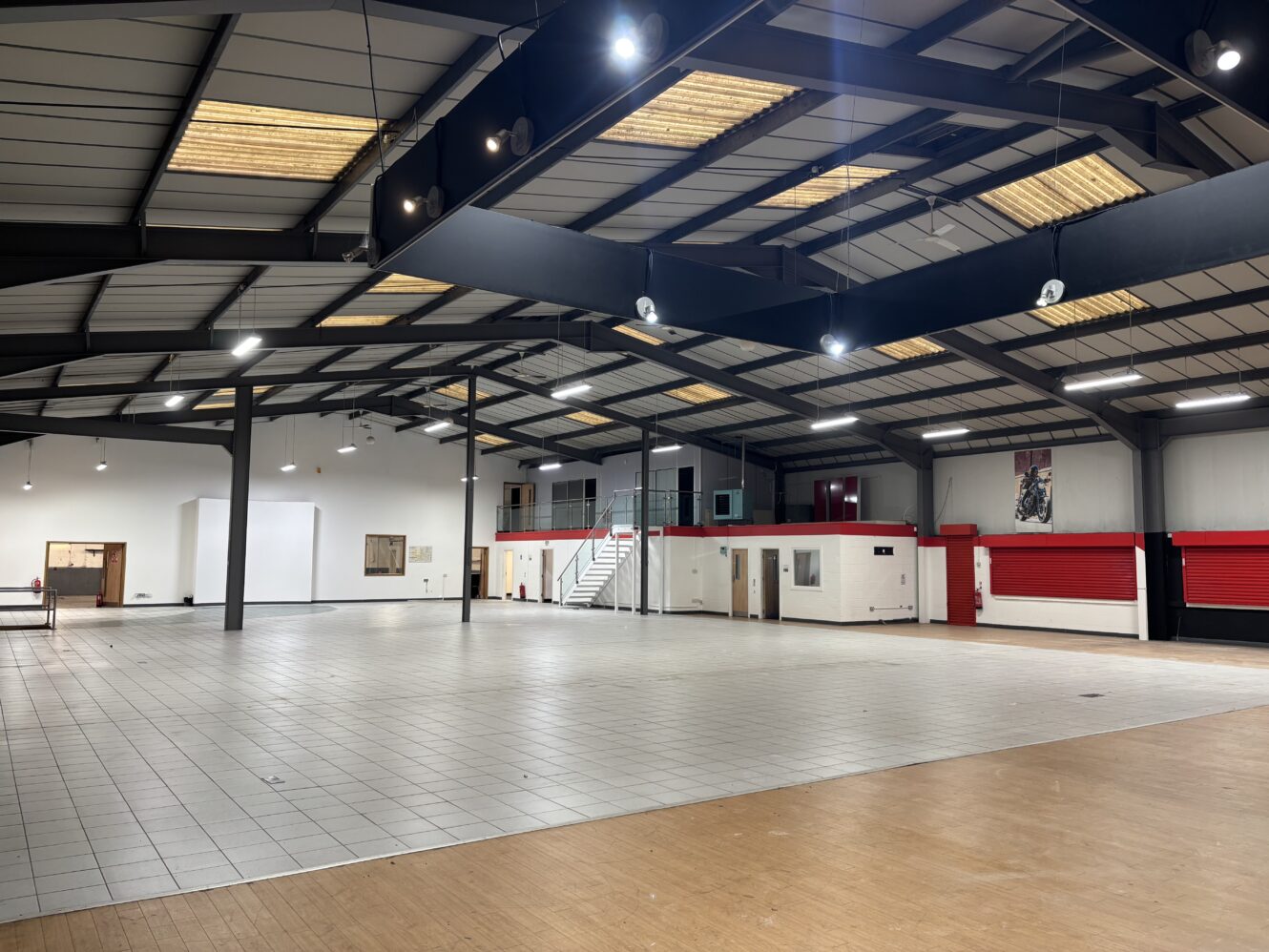 Spacious, empty warehouse interior with tiled flooring, exposed beams, fluorescent lighting, and a mezzanine level with stairs.