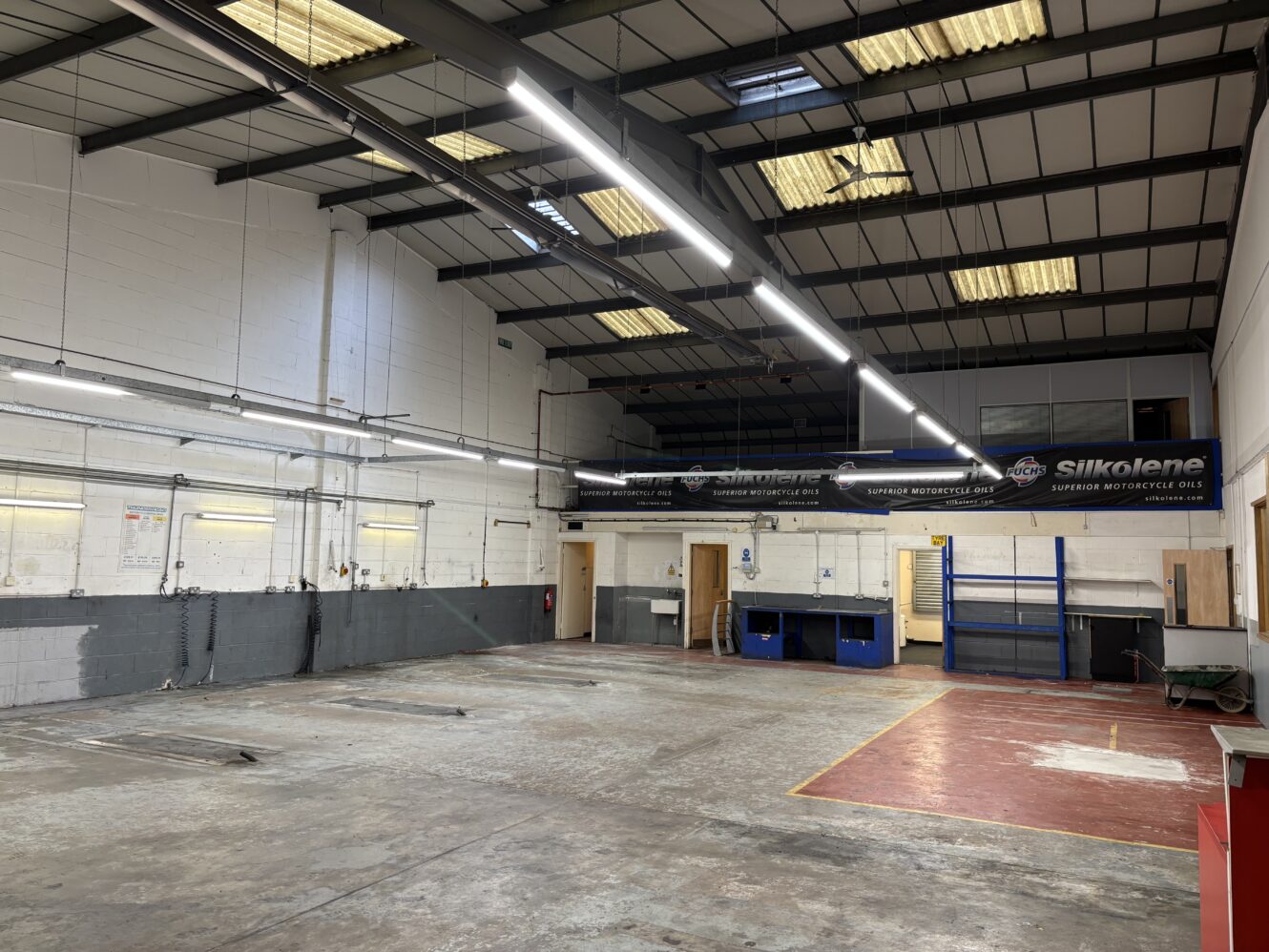 Large, empty industrial workshop with concrete floor, high ceiling, fluorescent lighting, workbenches, and storage shelves against white and gray walls.