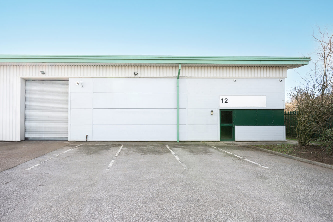 Empty parking lot in front of a light industrial building with a closed roller door, green trim, and the number 12 displayed on the wall.