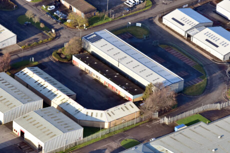 Aerial view of industrial buildings with metal roofs, parking lots, surrounding fencing, and nearby roads, captured during daylight.
