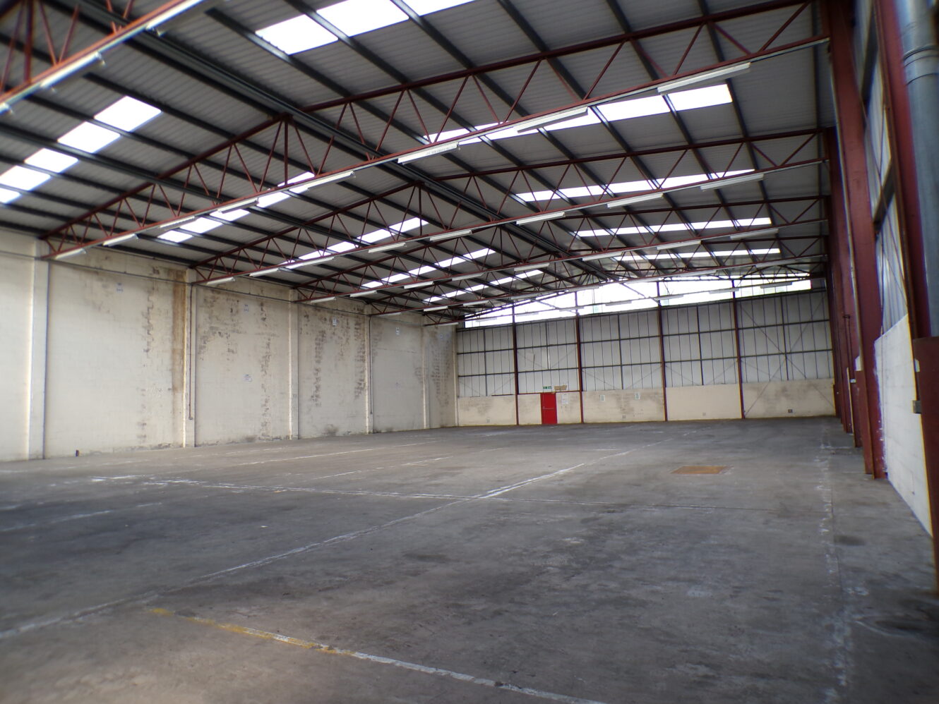 An empty industrial warehouse with high ceilings, exposed beams, concrete floor, and a red door on the far wall. Natural light enters through windows in the roof.
