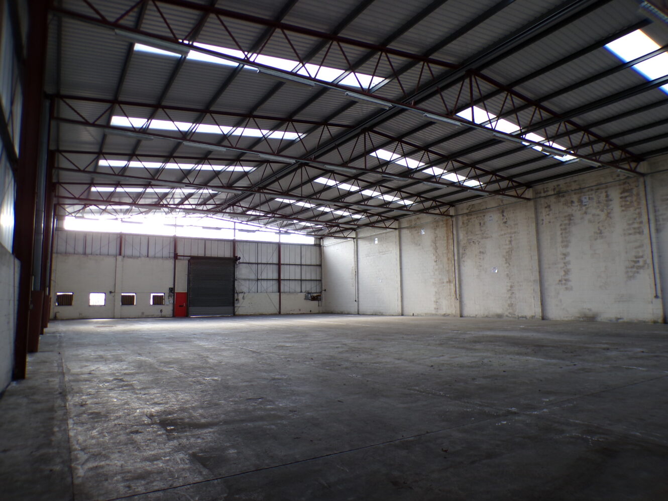 Interior of an empty warehouse with high ceilings, exposed beams, concrete floor, and natural light coming through windows and skylights.