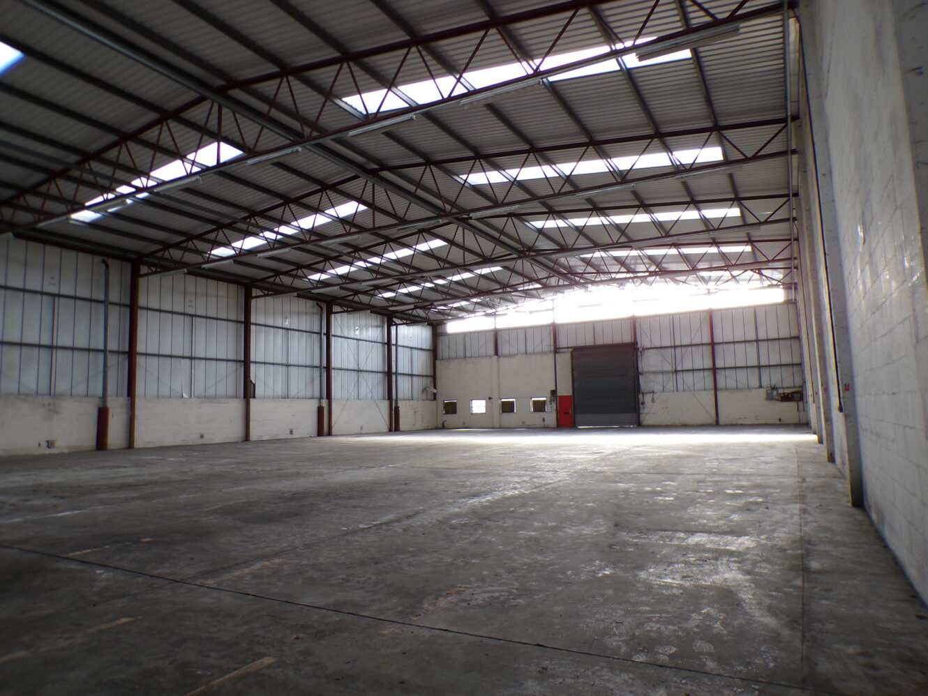Interior of an empty industrial warehouse with concrete floor, high metal ceiling, large windows, and a closed roller door at the far end.