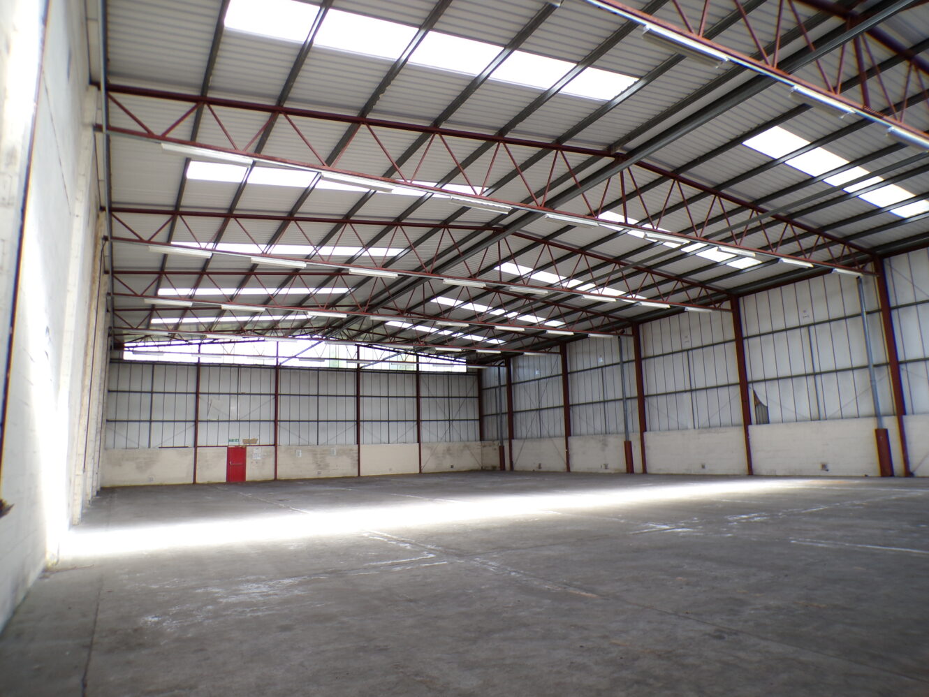 Empty warehouse interior with high ceilings, metal beams, concrete floor, and large windows letting in natural light.