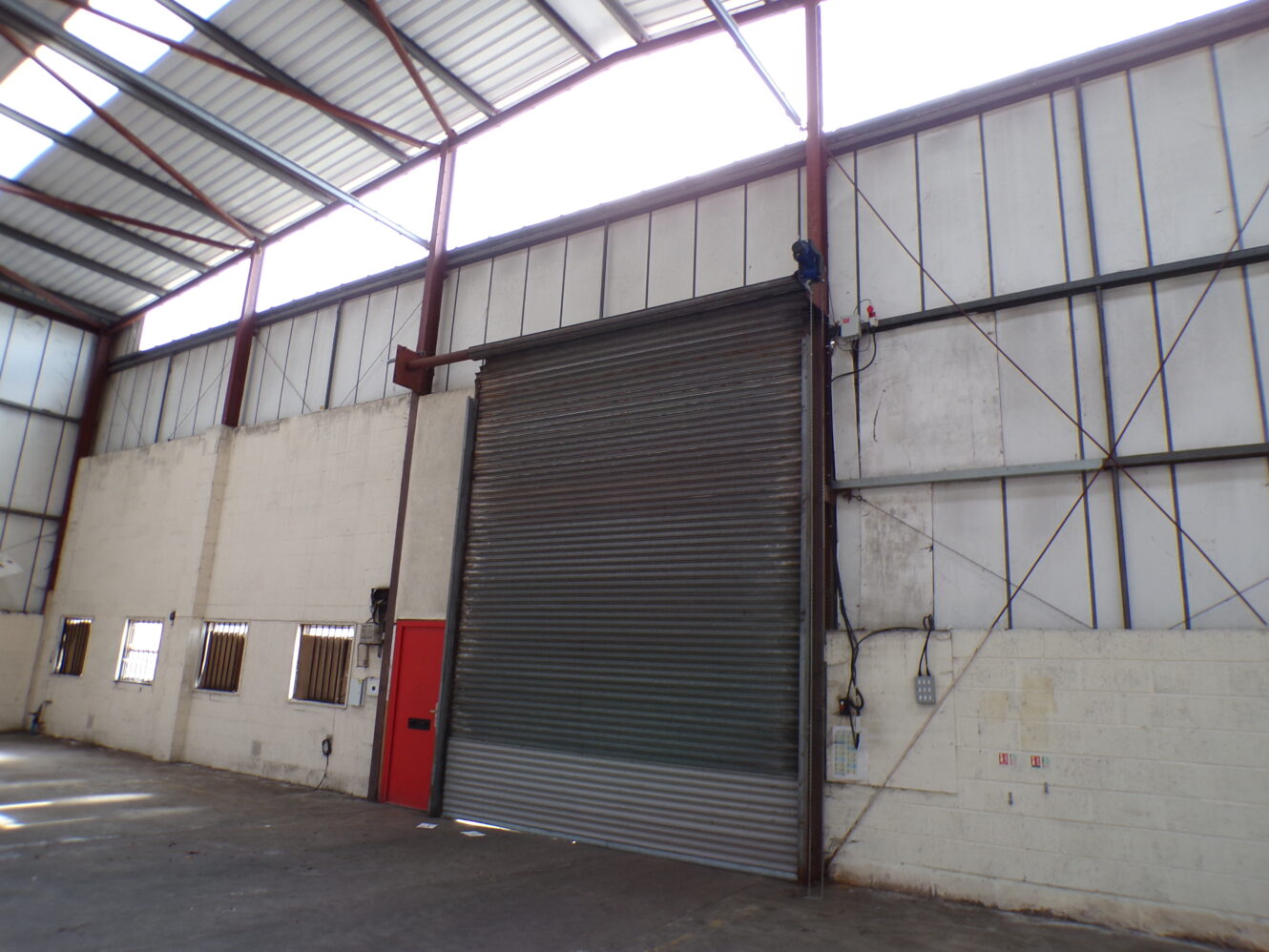 Large, partially open metal roll-up door in an empty warehouse with high ceilings, exposed beams, and a red personnel door on the left.