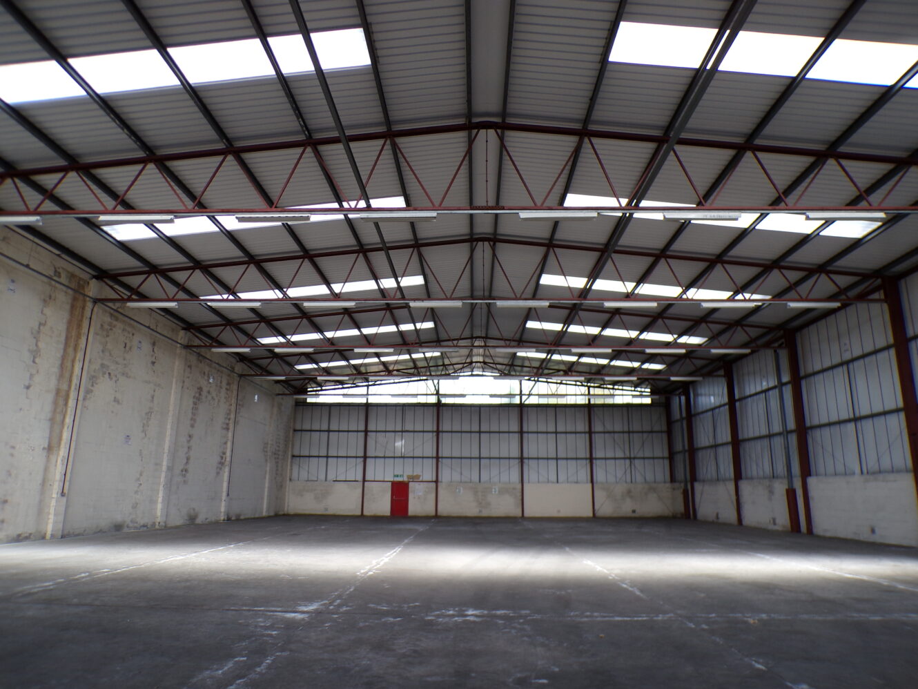 Interior of an empty warehouse with high ceilings, exposed beams, concrete floor, and red double doors at the far end. Skylights provide natural light.