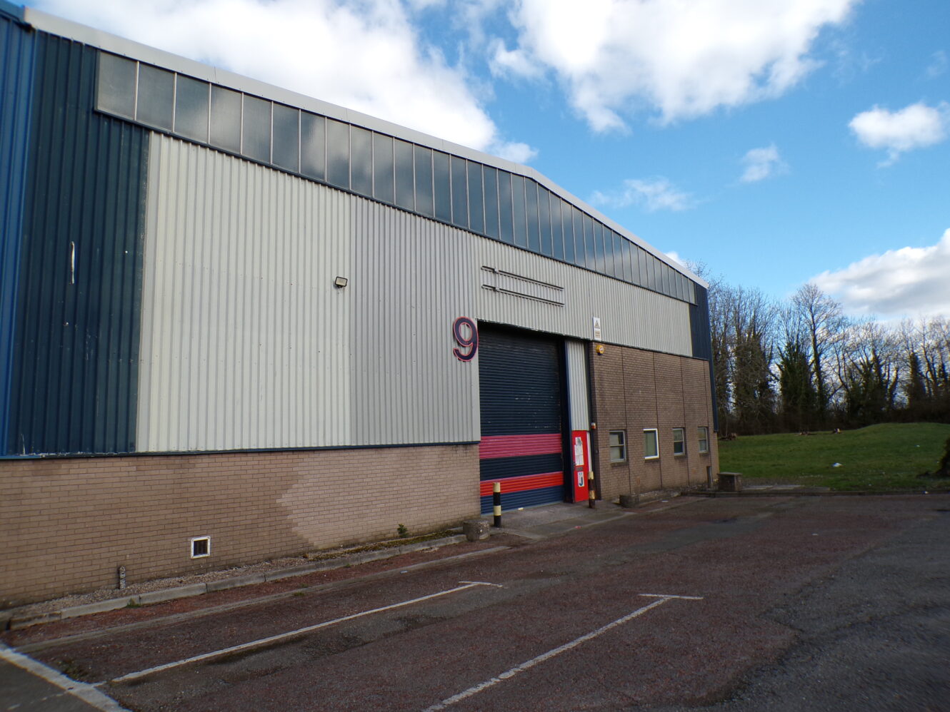 Warehouse building with a large blue and red roller door, marked with the number 9, adjacent to an empty parking lot and grassy area under a partly cloudy sky.