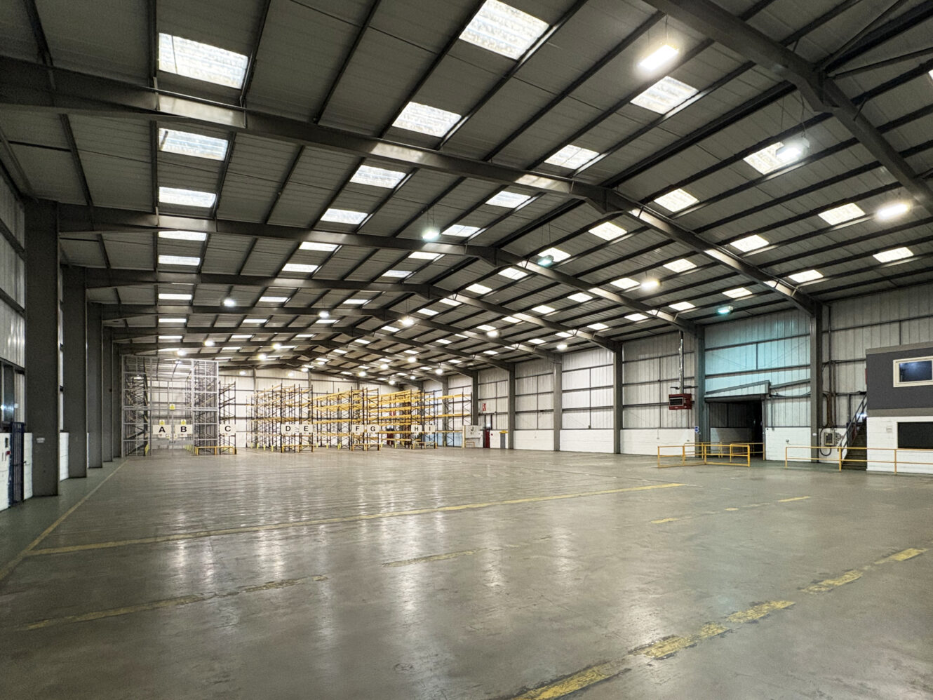 Interior of a spacious, empty warehouse with high ceilings, exposed beams, and shelves along the far wall. The space is clean with polished floors and overhead lighting.