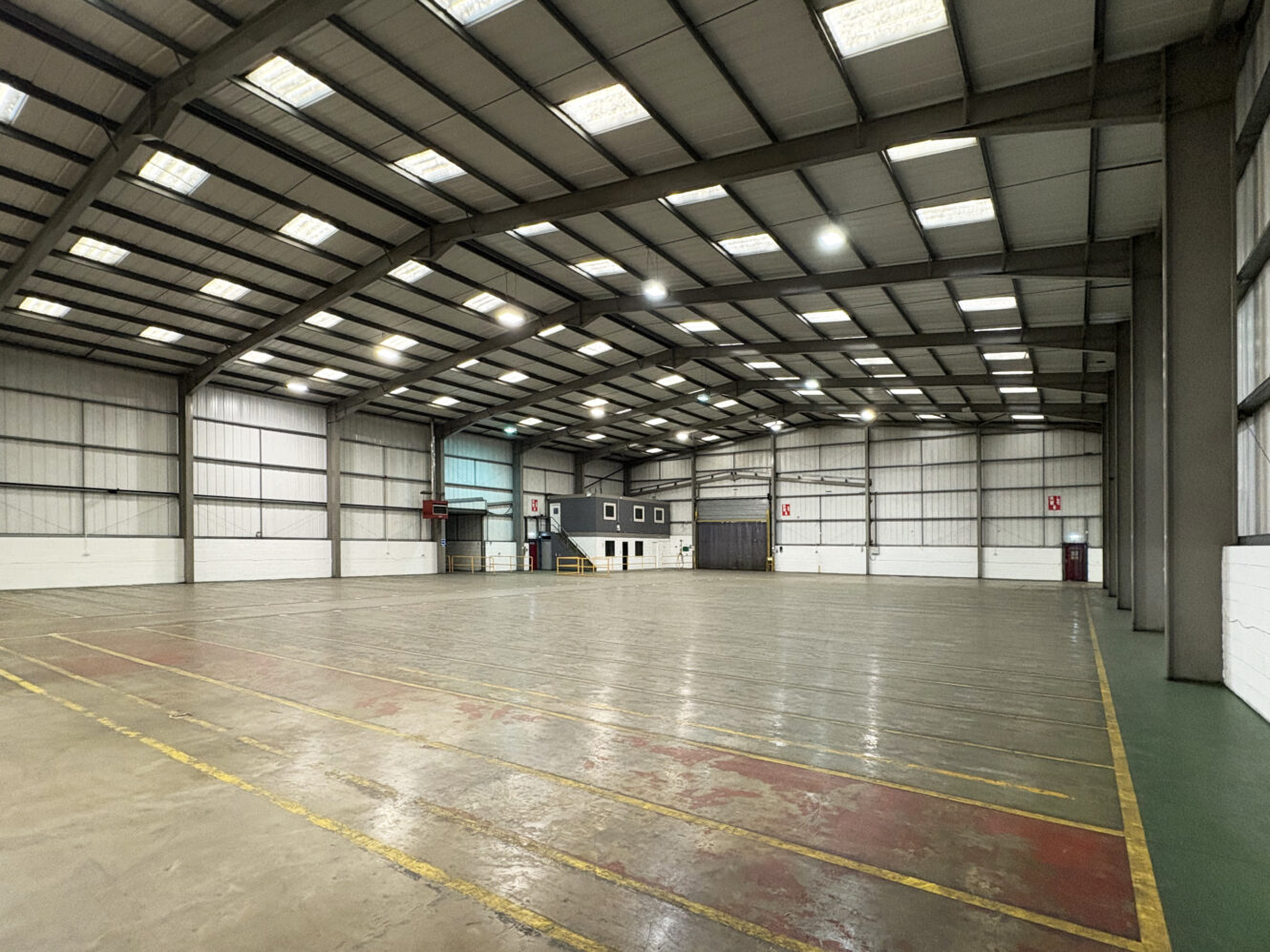 Spacious empty warehouse with high ceilings, overhead lights, concrete floor, and painted dividing lines; office area visible at the far end.