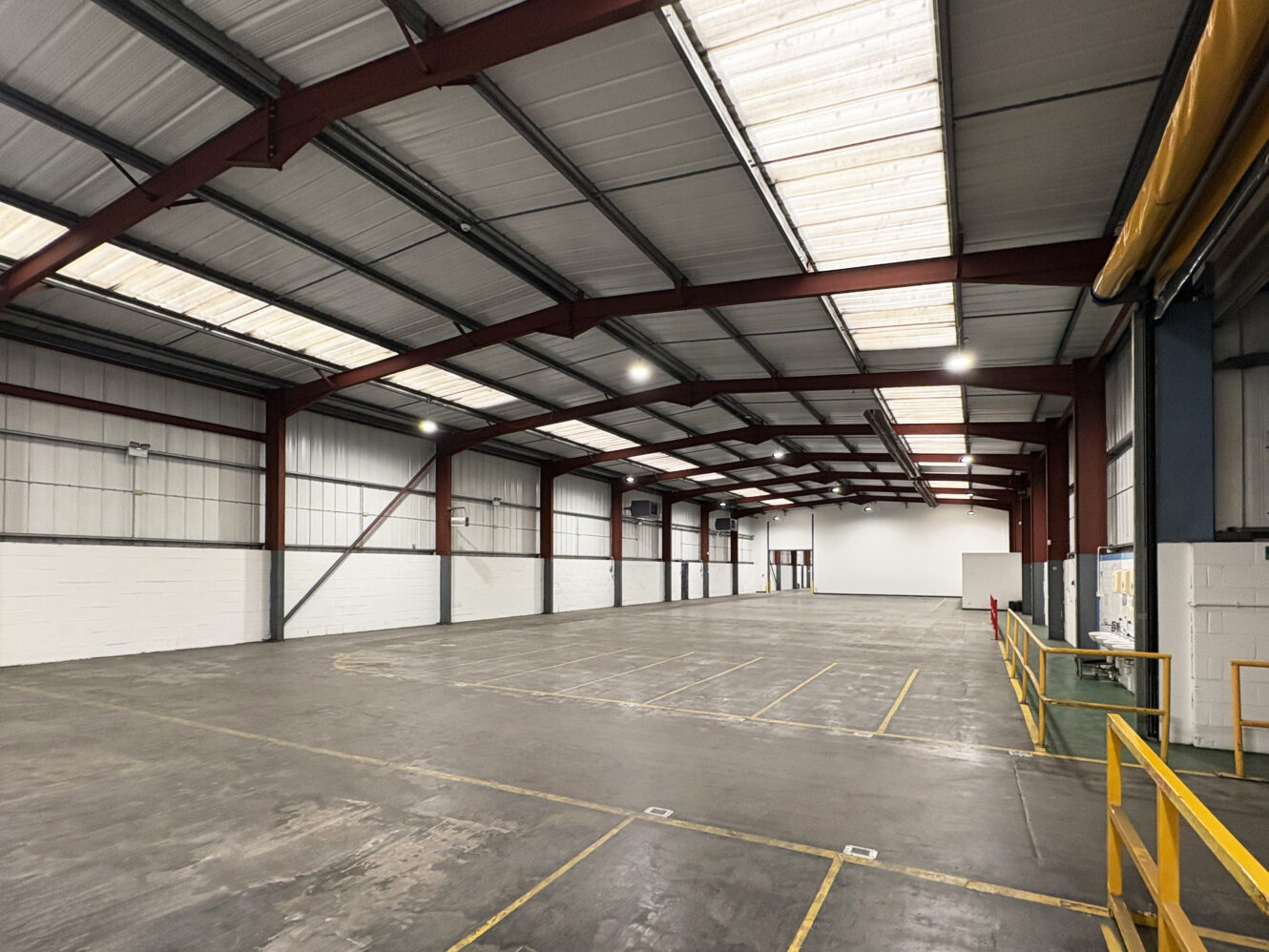 Spacious, empty warehouse interior with high ceilings, overhead lights, metal beams, and marked zones on the concrete floor.