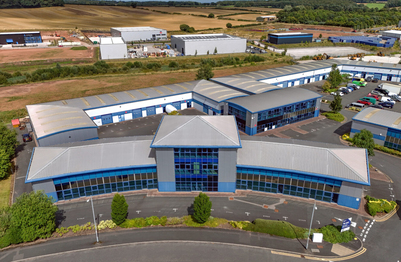 Aerial view of a modern industrial business park with multiple blue and gray warehouse-style buildings surrounded by paved roads and green landscaping.