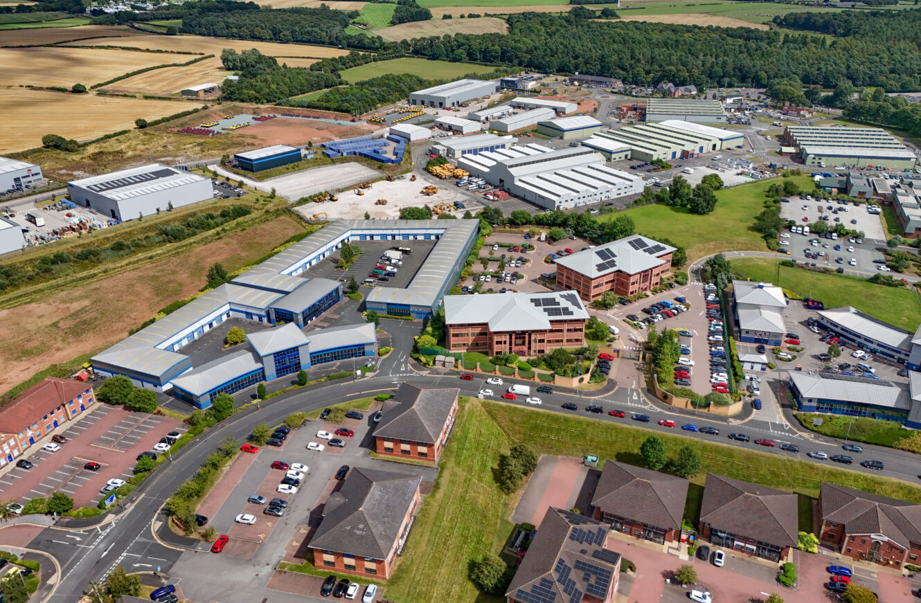 Aerial view of an industrial business park with large warehouses, office buildings, parking lots, cars, and surrounding fields and trees.