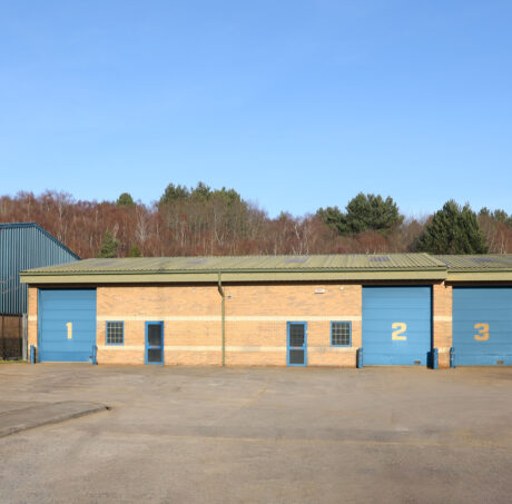 A row of three brick industrial units with blue doors numbered 1, 2, and 3, set against a backdrop of trees and clear blue sky.
