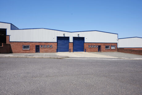 A large industrial warehouse with two blue roller doors and several small windows, set in a brick and metal building, under a clear blue sky.