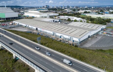 Aerial view of a large industrial warehouse with multiple loading bays, adjacent to a highway with several vehicles, and a cityscape in the background.