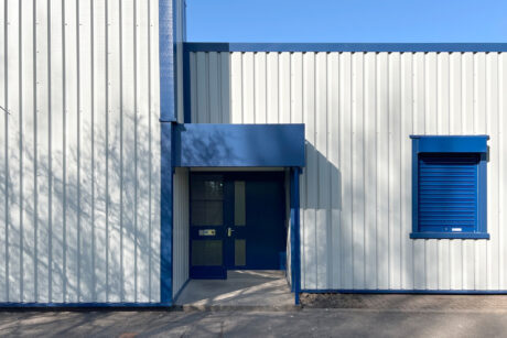 A modern industrial building with white corrugated metal walls, a blue door, and a blue shuttered window on a sunny day.