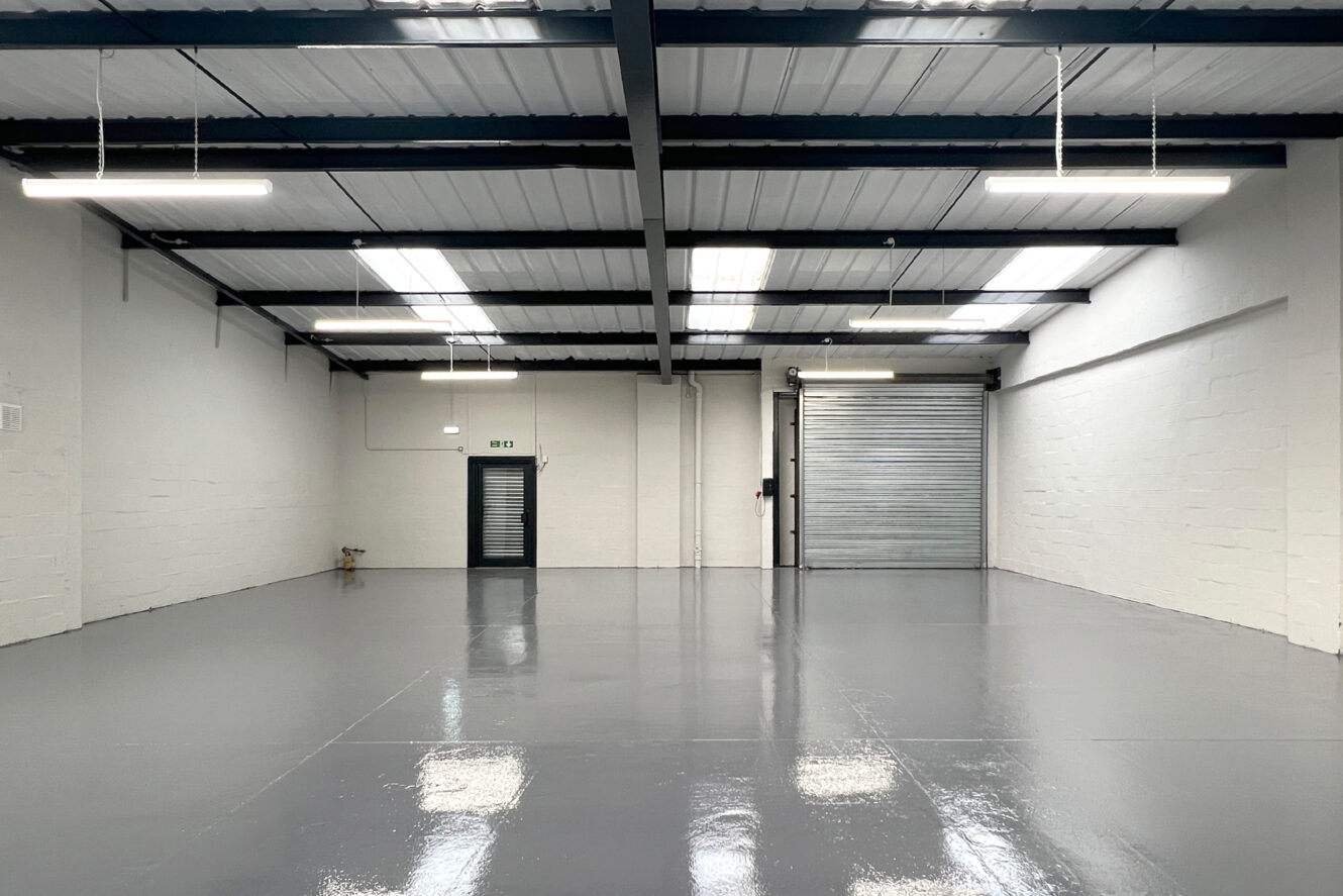 Empty industrial warehouse with glossy gray floor, white walls, exposed ceiling beams, fluorescent lights, a black door, and a large metal roll-up garage door.
