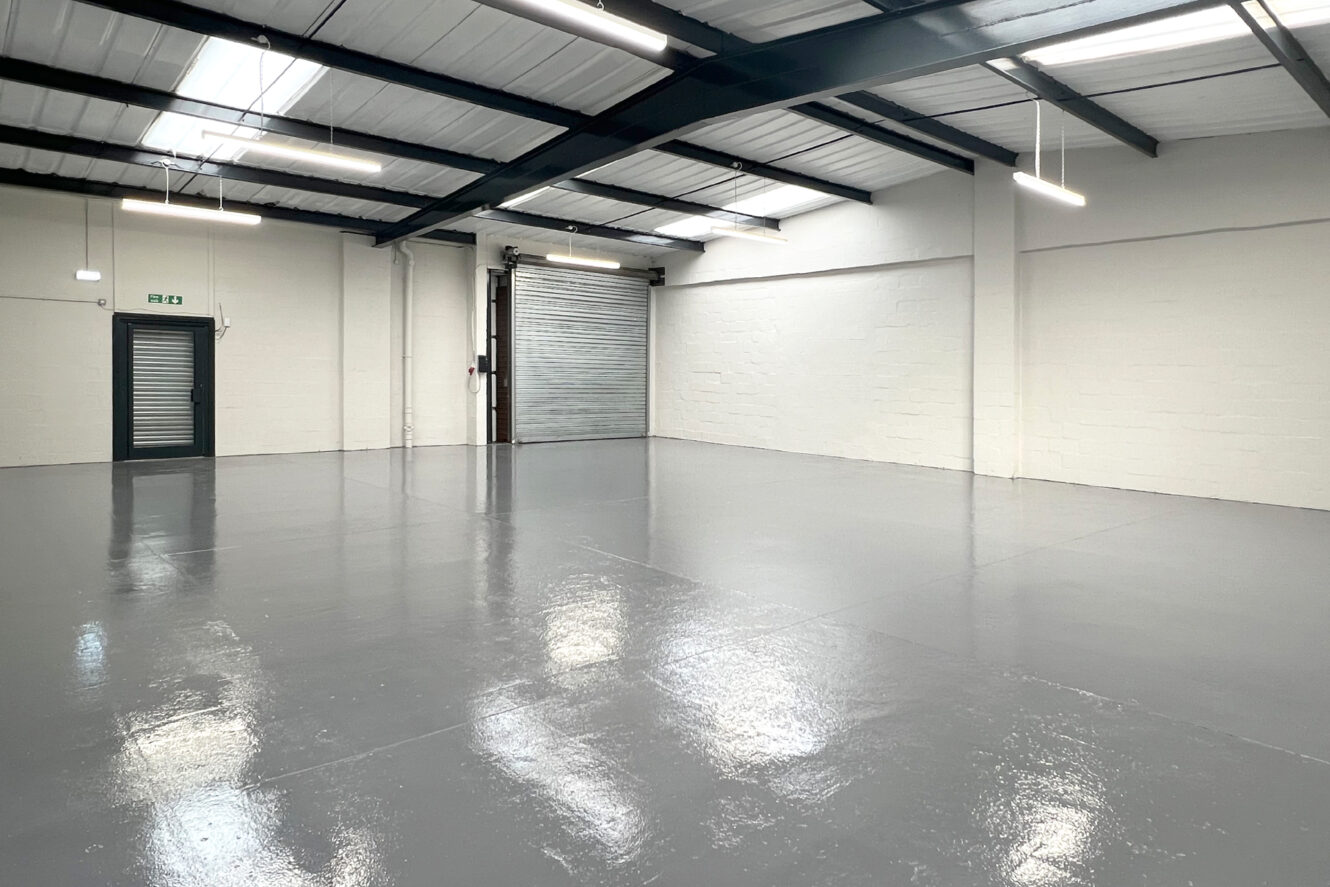 Empty industrial warehouse unit with polished grey floor, white walls, exposed beams, overhead lights, a metal shutter door, and an emergency exit.