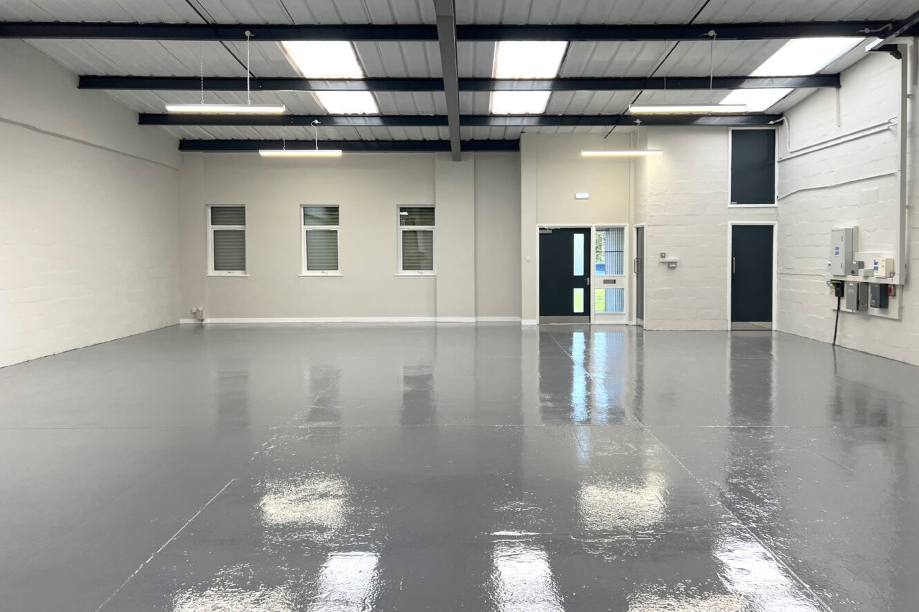 Large, empty industrial space with polished concrete floors, white walls, high ceilings, fluorescent lighting, windows, and doors along the back wall.